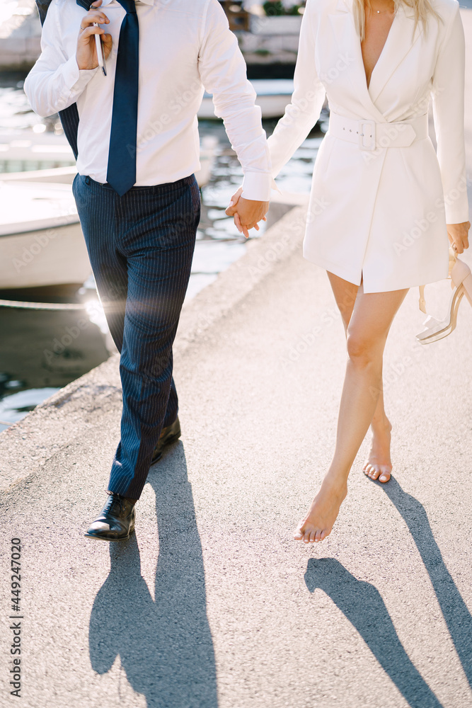 Groom with the barefoot bride walk holding hands along the pier on the background of yachts
