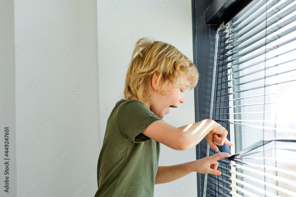 little joyous boy is trying to open jalousie, have fun, smile. side view portrait. caucasian blonde kid boy in green t-shirt alone at home, standing next to window little joyous boy is trying to open jalousie, have fun, smile. side view portrait. caucasian blonde kid boy in green t-shirt alone at home, standing next to window