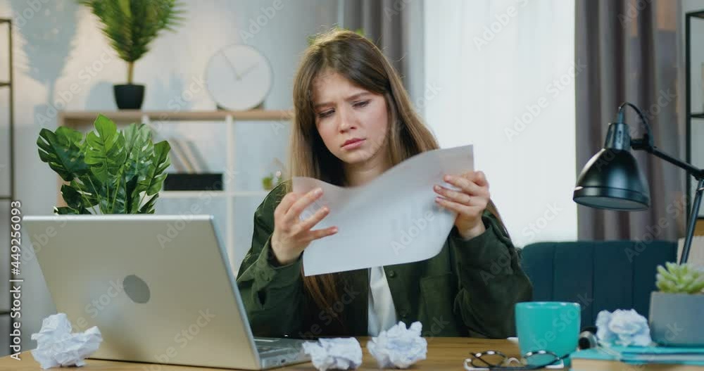 Pretty unhappy stressed young female freelancer working on laptop and checking datas from report with information on screen,crumpling paper and throwing it to pile of crumpled papers on the table Pretty unhappy stressed young female freelancer working on laptop and checking datas from report with information on screen,crumpling paper and throwing it to pile of crumpled papers on the table