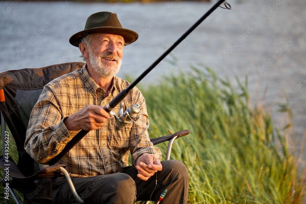 Fishing is very common hobby in coastal areas areas with rivers. Senior caucasian Man fishing in lake alone, in countryside, holding Fishing rod in hands, in casual wear