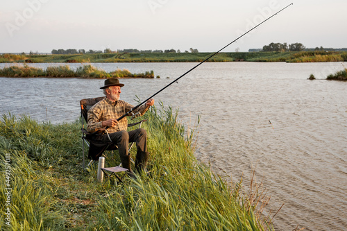 Portrait Of Senior Caucasian Man On Camping Holiday With Fishing Rod, Gra-haired Sit On Chair Alone Outdoors In Countryside Nature. Relax Leisure Time