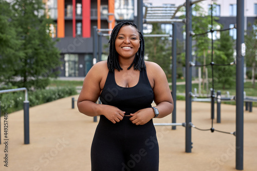 Positive afro american woman in black tracksuit posing at camera outdoors  smiling. Obese overweight lady enjoy sport on fresh air  happy to be healthy and sporty. weight loss  people lifestyle