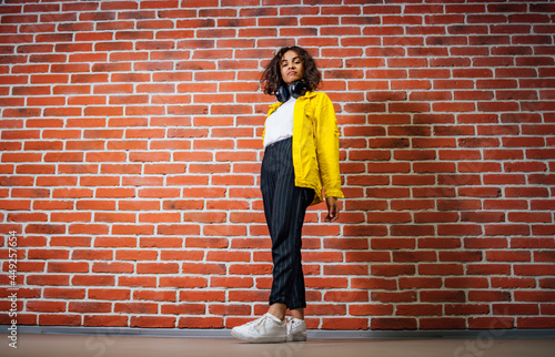 Full length view of the beautiful young sexy girl in a yellow jacket listening to music in headphones on a brick wall background. Excited woman looking at the camera. Positive emotions concept