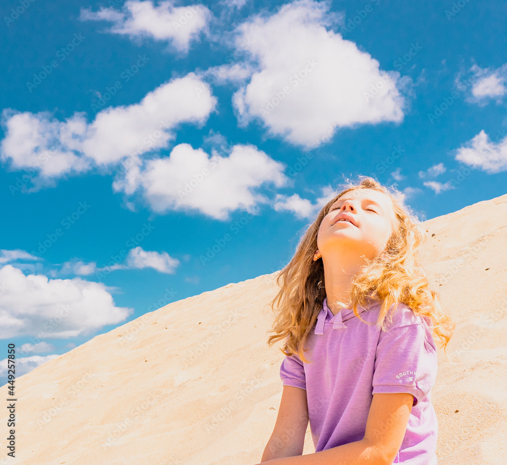 Young cute girl in hat sitting on the sand beach. Fashion model girl posing on the sand dunes. Blue summer sky as background. Happy childhood. High quality photo