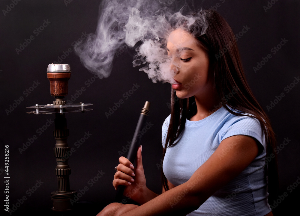 Obraz premium The girl smokes a hookah on a black background. Smoke comes out of the mouth. The pleasure of smoking