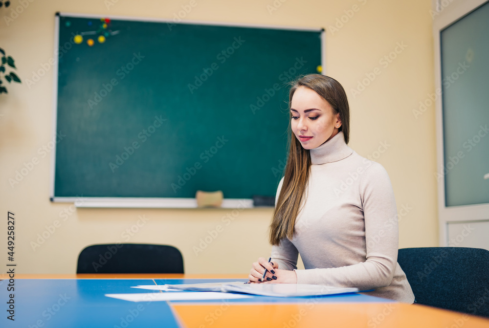 Beautiful young teacher checking notes while sitting at the table. Woman teacher in classroom. Blackboard background Beautiful young teacher checking notes while sitting at the table. Woman teacher in classroom. Blackboard background