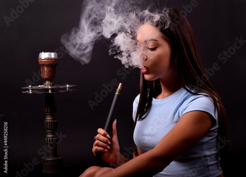 The girl smokes a hookah on a black background. Smoke comes out of the mouth. The pleasure of smoking
