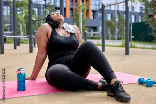 Tired fat afro female having rest on fitness mat after workout, take a break with bottle of water and towel, in sports ground, throwing head back. attractive mixed race lady engaged in fitness