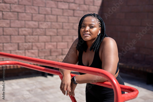 Dreamy african woman in black sportive clothes having rest, looking at camera in city, in sports ground, relaxing, thinking, having some minutes to take a break during jogging or fitness workout