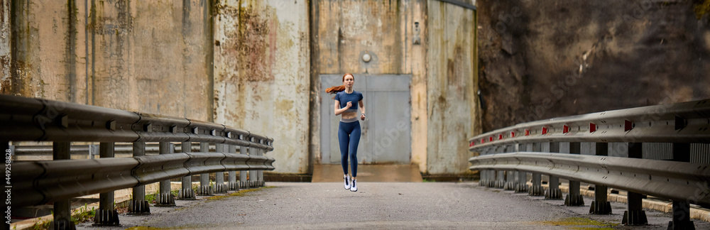 view from distance on young slim sportswoman running on bridge alone outdoors, in sportive clothes, looking confident and focused, leading healthy lifestyle, side view, copy space. sport and fitness
