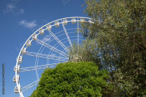 Large white Ferris wheel against the blue bright sky, behind the trees, bottom view. Concept entertainment. © RinaChu