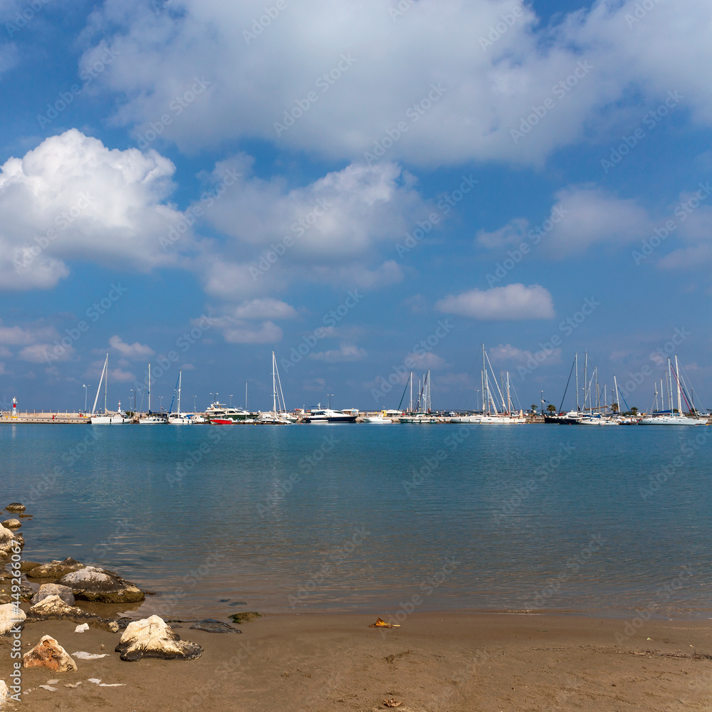 view from a rocky wild beach to white yachts moored at the pier in the Greek town of Rethymnon against a background of blue sky