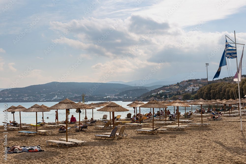 sandy beach with sun loungers and reed umbrellas and vacationers against the backdrop of the sea, mountains and blue sky. Greece. Agios Nikolaos sandy beach with sun loungers and reed umbrellas and vacationers against the backdrop of the sea, mountains and blue sky. Greece. Agios Nikolaos