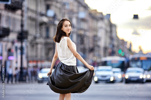 Slender teen girl in long skirt in front of busy road traffic on city street an evening.