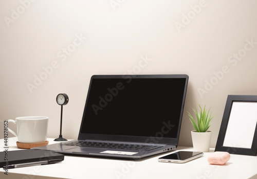 Picture of laptop on a white table with a mug a clock a small bush a notebook a phone a picture frame and a headphone case