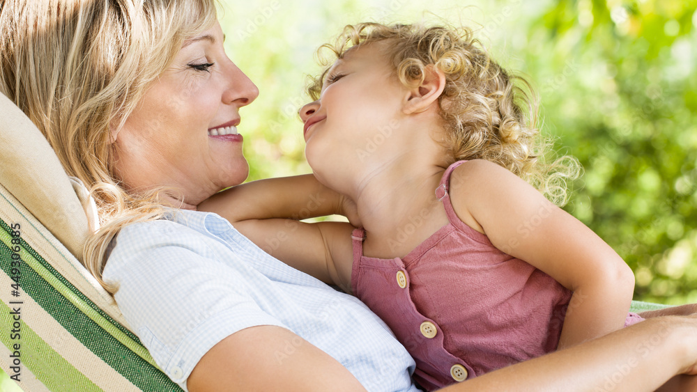 happy and smiling mom hugs her little girl daughter child blue eyes with blond curly hair, together lying on the hammock in the green home garden, lovely hugs and caresses of family concept happy and smiling mom hugs her little girl daughter child blue eyes with blond curly hair, together lying on the hammock in the green home garden, lovely hugs and caresses of family concept