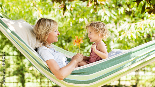 smiling mom and little girl daughter child blue eyes with blond curly hair, together lying on the hammock in the green home garden, plays with lily flower, happy family and spring time concept photo