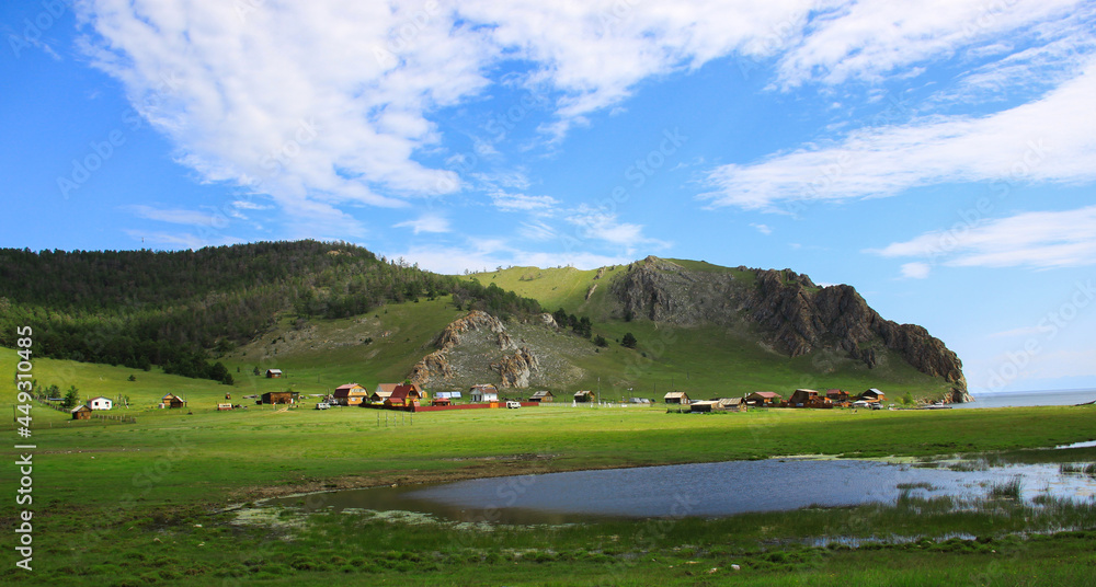 A bright summer landscape. The small Uzura village is located at the foot of the mountain. A bright green meadow is located on the hillsides under a blue sky with white clouds. A coast of Lake Baikal