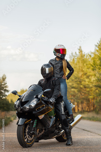 Motorcyclist in leather jacket and helmet sits on sports motorcycle, girl stands, blurred background, copy space