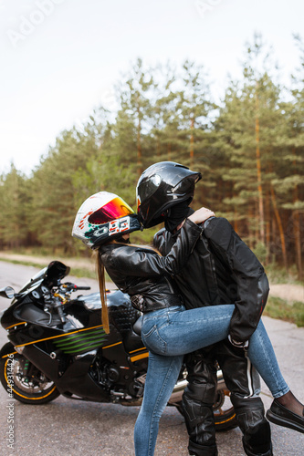 Motorcyclist in leather jacket and helmet stands next to sports motorcycle and hugged girl in helmet and tilted her  blurred background  copy space