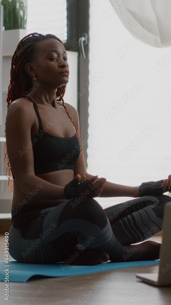 Slim black woman sitting in lotus pose on yoga map with closed eyes meditating on floor looking at laptop computer. Athlete adult relaxing during aerobic class doing breathing exercises