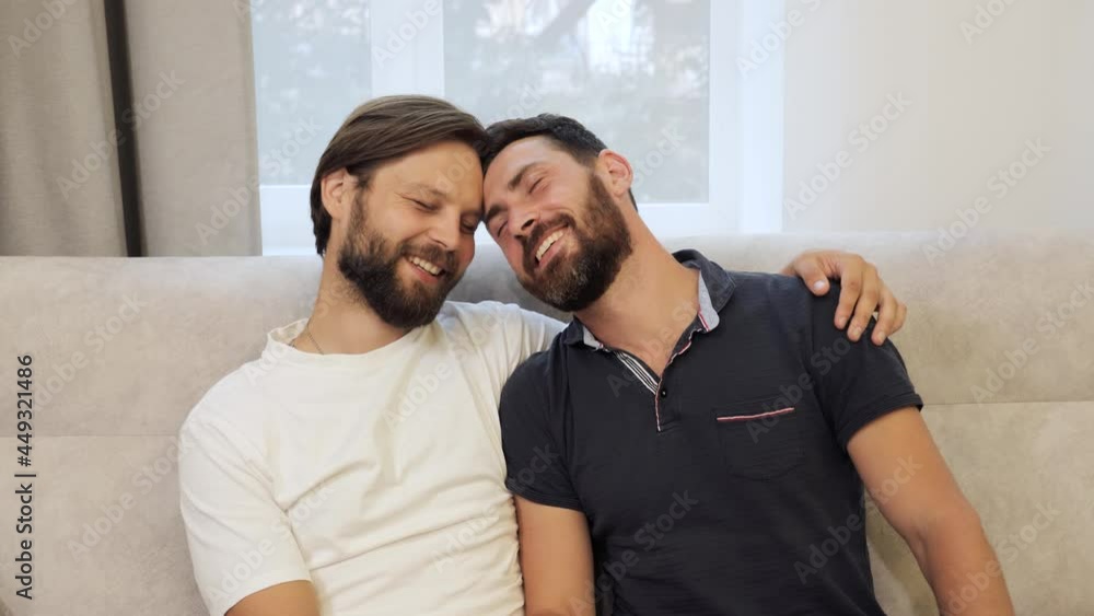 Portrait of two affectionate gay men in love wearing casual clothes hugging smiling to camera sitting on the couch. Homosexual couple dating at home. LGBTQI, Pride Event, LGBT Pride Month. Portrait of two affectionate gay men in love wearing casual clothes hugging smiling to camera sitting on the couch. Homosexual couple dating at home. LGBTQI, Pride Event, LGBT Pride Month.