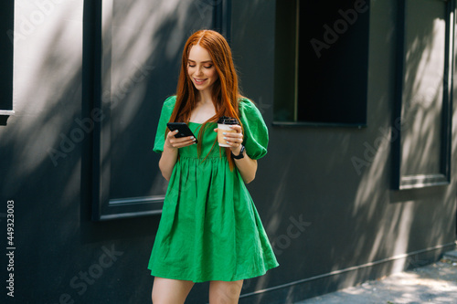 Happy redhead young woman wearing green dress walking at city street in sunny summer day, holding takeaway coffee cup, using mobile phone. Smiling attractive lady browsing smartphone outdoors. © dikushin