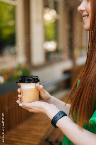 Vertical cropped shot of smiling redhead young woman holding cup with hot coffee sitting at table in outdoor cafe terrace in summer day, blurred background, selective focus, close-up.