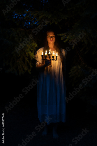 A young woman in the dark woods holding a candelabrum. Wearing a long white dress.