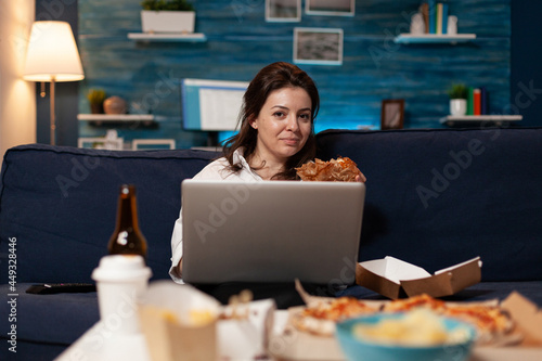 Caucasian female sitting on sofa eating tasty delicious burger while working on laptop computer in living room late at night. Woman enjoying takeaway food home delivered. Fastfood lunch meal order