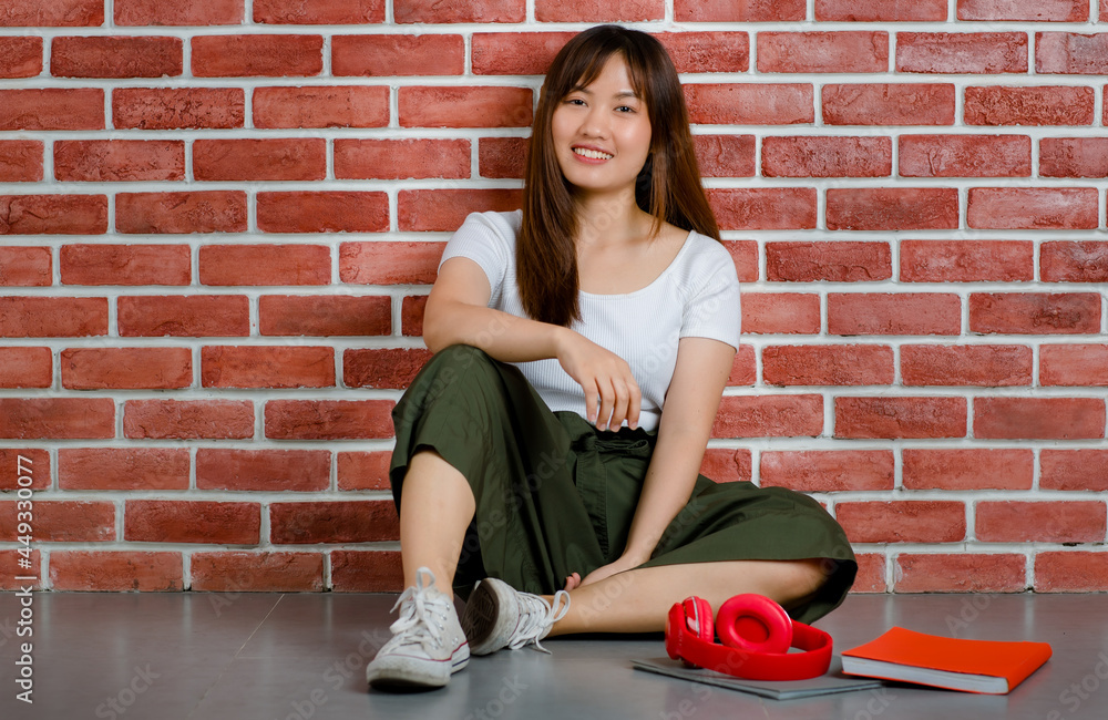Full length of young attractive Asian woman in white t-shirt and green skirt sitting on the floor with books and red headphone on the floor against orange brick wall background