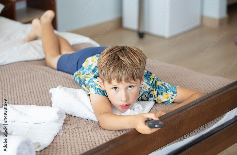 Fototapeta premium The child lies on the bed with the TV remote control.