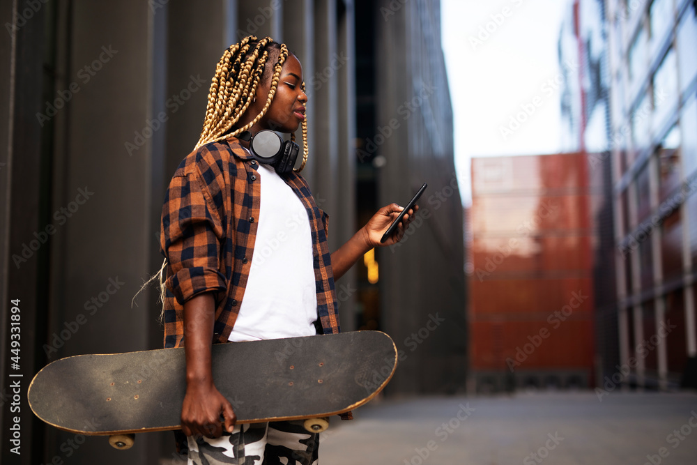 Obraz premium Portrait of modern african girl with skateboard. Young stylish woman using the phone.