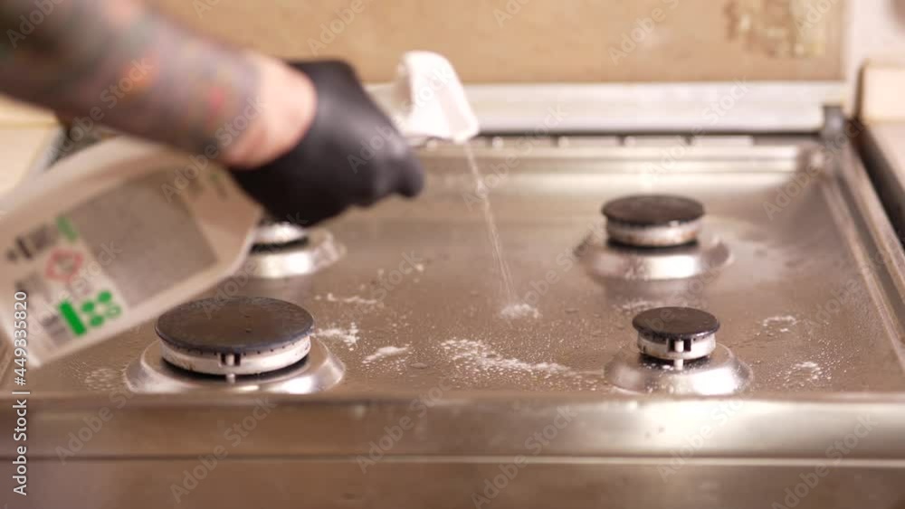 A man in the kitchen washes the gas stove, hands in rubber gloves with the help of special cleaning agents and a sponge will remove dirt from the surface of the gas cooking stove. A man in the kitchen washes the gas stove, hands in rubber gloves with the help of special cleaning agents and a sponge will remove dirt from the surface of the gas cooking stove.