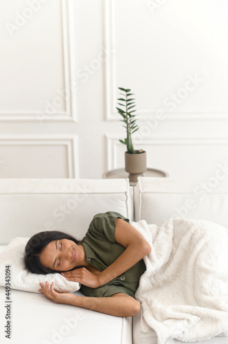 Happy beautiful Indian woman taking a nap on the sofa at home, serene mixed-race female sleeping, rested on the pillow, covered with blanket, vertical shot