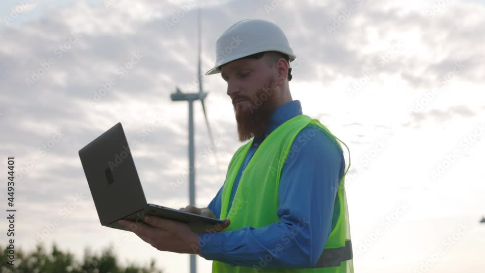 Male technician employee in safety vest and white industrial helmet browsing computer laptop technology to control wind turbine system. Windmill worker. Male technician employee in safety vest and white industrial helmet browsing computer laptop technology to control wind turbine system. Windmill worker.