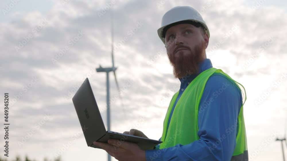 Bearded young caucasian engineer programming on laptop near wind turbines. Industrial worker in protective suit at work. Wind farm. Sustainable energy. Bearded young caucasian engineer programming on laptop near wind turbines. Industrial worker in protective suit at work. Wind farm. Sustainable energy.