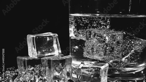 A transparent glass beaker with a colorless liquid ice cubes floating in it and scattered on the black surface on which the glass stands