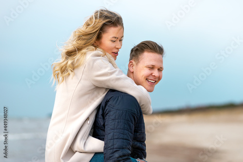 Happy man giving piggyback ride to his woman and laughing at beach. Smiling guy in love carrying on back her girlfriend.