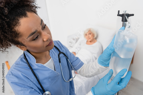 African american nurse in latex gloves holding bottle of intravenous therapy near blurred patient in hospital ward