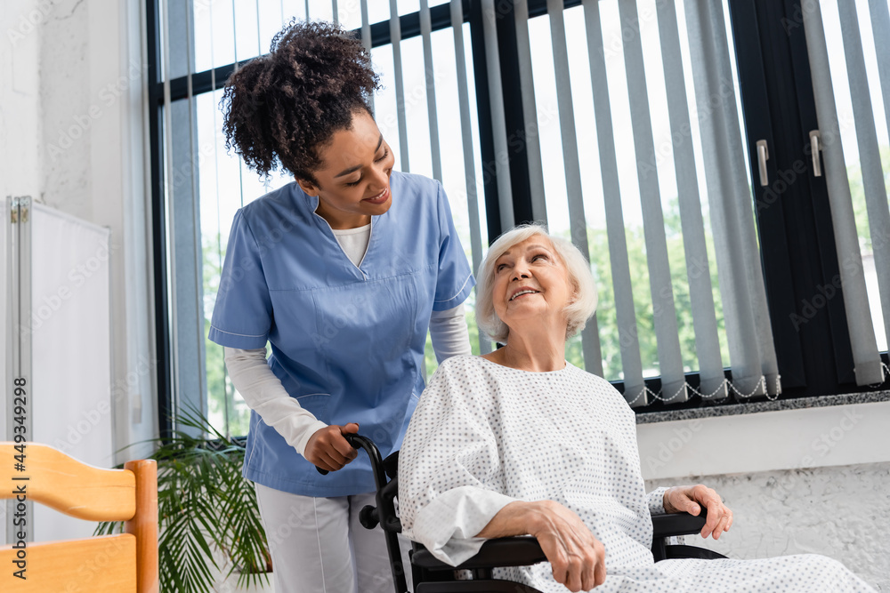 Smiling african american nurse looking at senior patient in wheelchair in hospital
