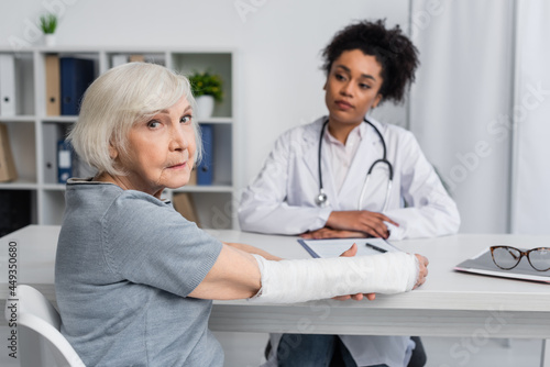 Senior woman with plaster bandage on arm looking at camera near blurred african american doctor