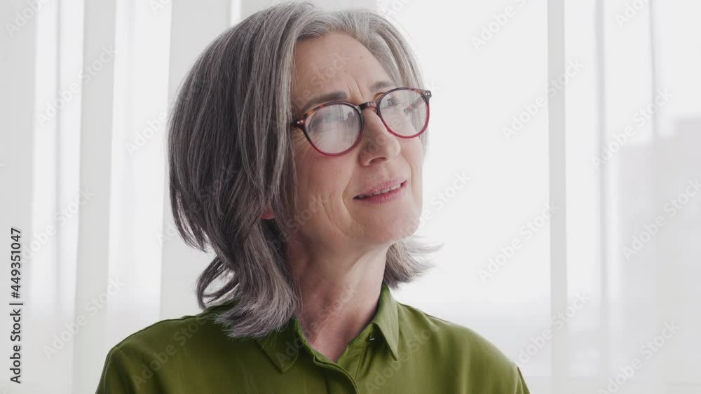 Attractive mature grayhaired lady dreaming standing near big windows of new flat Attractive mature grayhaired lady dreaming standing near big windows of new flat