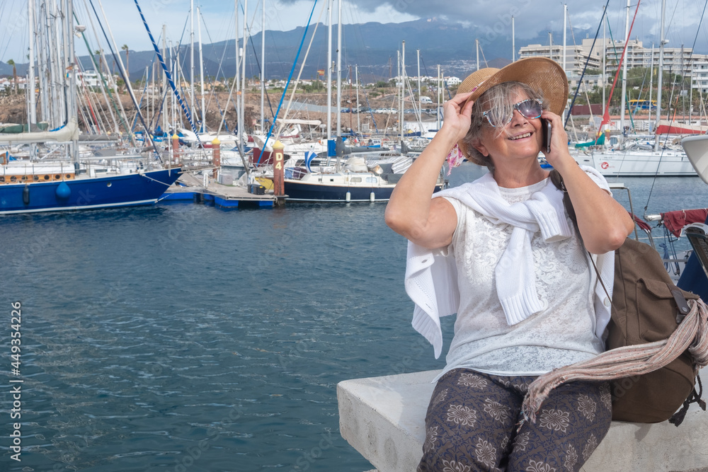 Smiling senior woman enjoying sea excursion at harbour sitting and using mobile phone