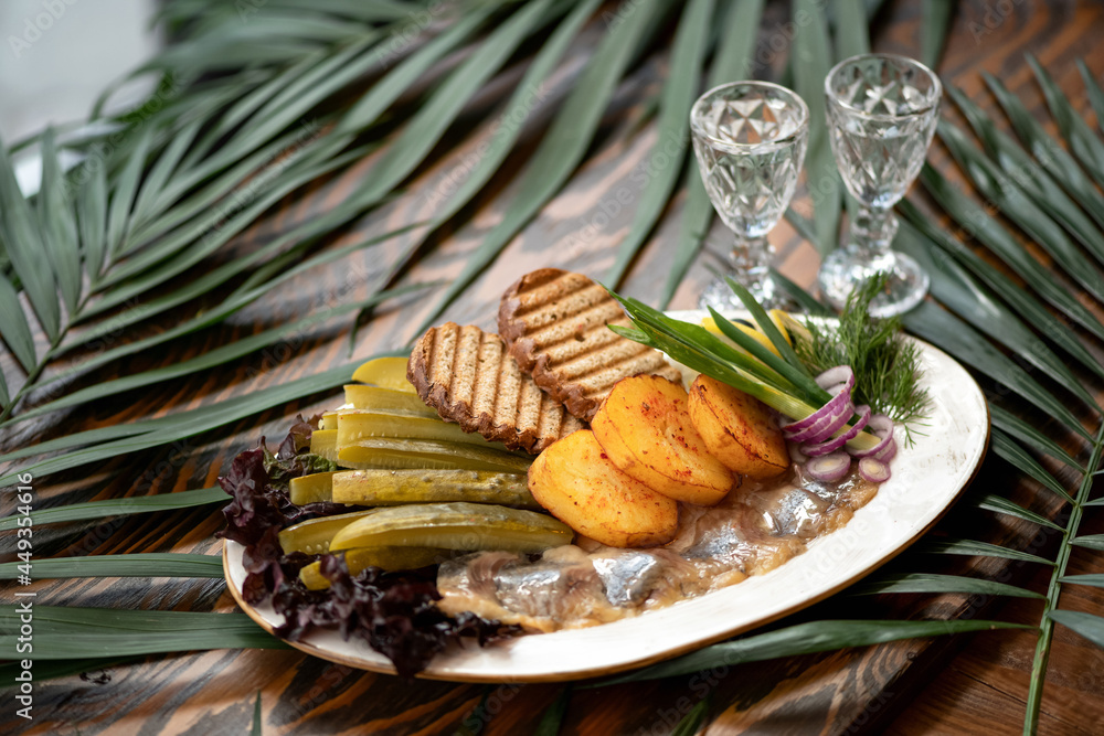 Festive Snack. Two glasses of alcohol shot or vodka on table decorated with palm leaves. Grilled bread, baked potatoes, herring, pickled cucumbers and greens on platter. Horizontal format. Soft focus. Festive Snack. Two glasses of alcohol shot or vodka on table decorated with palm leaves. Grilled bread, baked potatoes, herring, pickled cucumbers and greens on platter. Horizontal format. Soft focus.