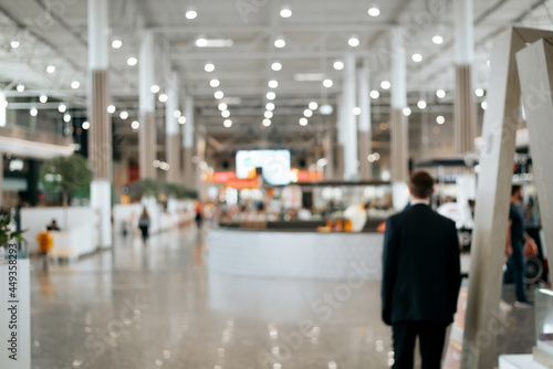 Defocused shopping mall background, large illuminated hall, security guard rear view. Blurred backdrop of a commercial space with people