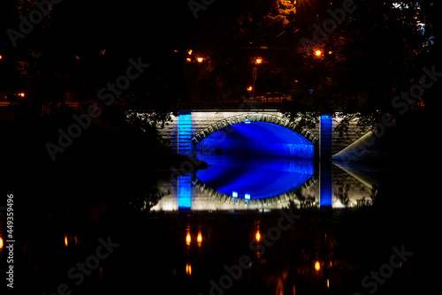 Night photo of a pedestrian bridge over a canal. The bridge is illuminated with blue lights that reflect in the water. Riga, Latvia