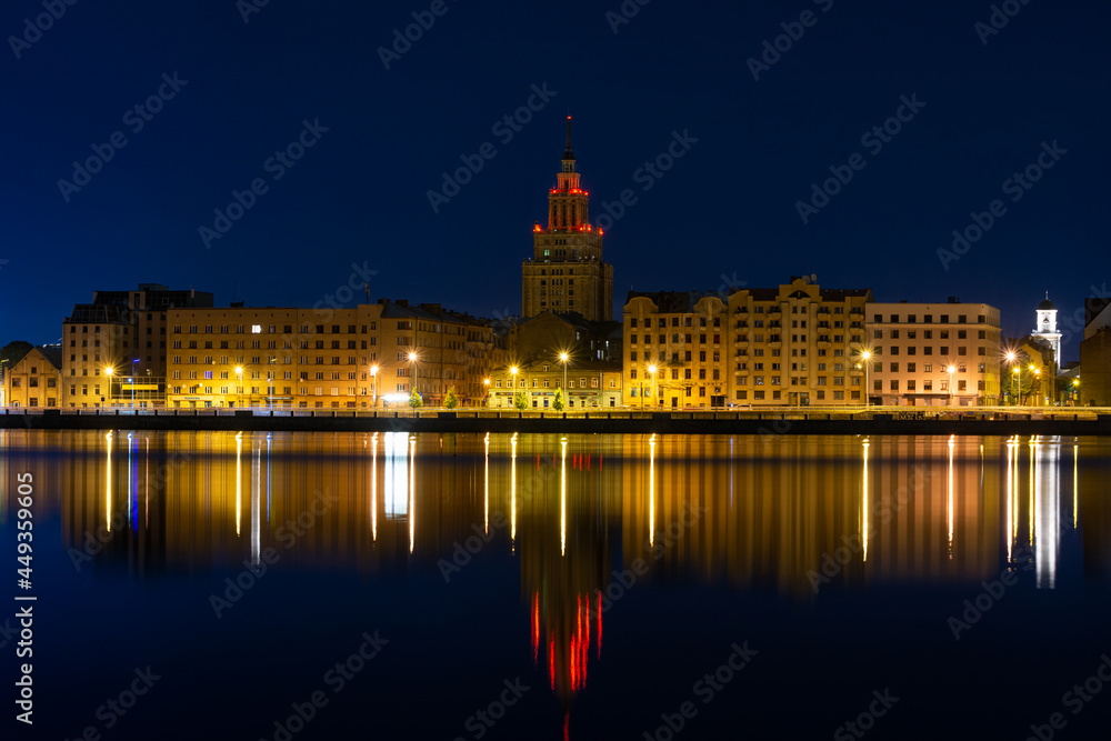 Summer dawn over the illuminated old buildings of the Moscow district and the quiet waters of the Daugava river in Riga, Latvia. Tower of the Science Academy building on the background.