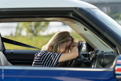 Tired exhausted woman of middle age lying on steering wheel in car driving. Unhappy depressed sleepy female try to stay awake after long road trip or bored in heavy traffic jam. Girl driver in vehicle