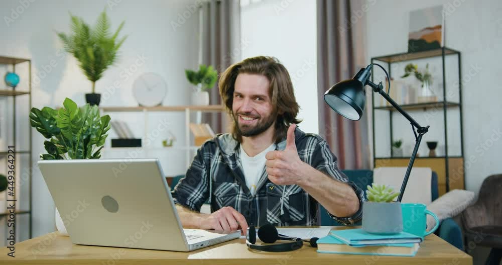 Portrait of good-looking smiling confident modern young bearded man in casual clothes which showing sign thumb up into camera in home office Portrait of good-looking smiling confident modern young bearded man in casual clothes which showing sign thumb up into camera in home office